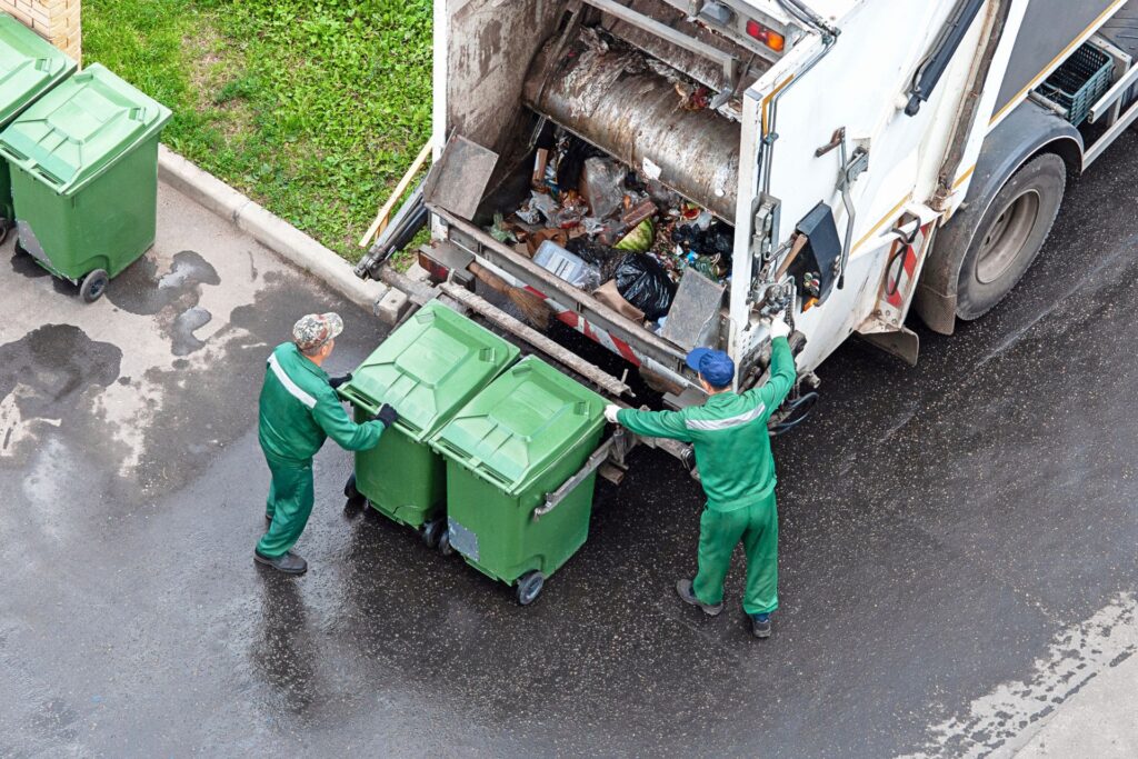 Two garbage collectors loading two green bins into their garbage truck